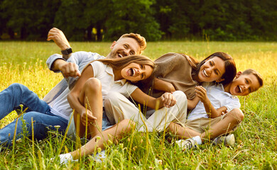 Happy young family of four smiling and having fun while sitting on green grass in park during summer rest. Laughing mother and father hugging their kids boy and girl lying on the field.