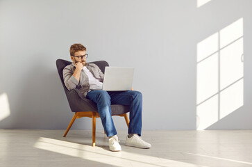 Pensive smart clever young man coder in casual shirt, jeans and glasses sitting in comfortable...