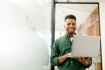Motivated Indian businessman stands in contemporary office hallway, using his laptop and beaming with cheerful smile looks at camera, male office employee, freelancer full of ideas and creativity