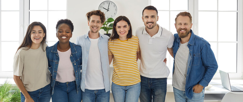 Happy diverse friends or coworkers. Banner with a group portrait of cheerful young multiracial people standing together, hugging each other, looking at the camera and smiling
