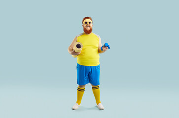 Fototapeta na wymiar Full-size studio shot of fat funny guy with yellow and blue fitness suit. Cheerful happy young man in sunglasses holds yellow sports mat and small dumbbells on isolated soft blue background.