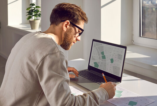 Cadastral engineer keeps records of land plots by making marks on map. Young engineer with cadastral plans and projects is sitting at desk with laptop in office. Real estate object, ownership rights.