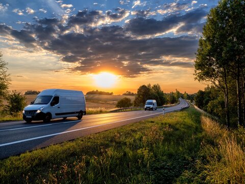 Two white fast delivery vans driving on the asphalt road in rural landscape at sunset with dark cloud