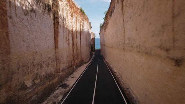 Aerial drone flying over asphalt road and high rock mountain walls on both sides. Bali Pandawa beach approaching between sandstone cliff, water and blue sky. Nature and travel concept