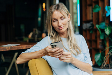 Young blonde modern woman sitting in a coffee shop uses a mobile phone to surf the internet.