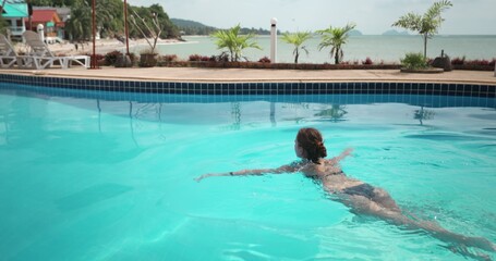 Woman swimming in pool with blue clear water in warm summer day. Young girl relaxing during refreshing in basin. Concept of summer vacation on tropical island, travel, tourism, recreation. Slow motion
