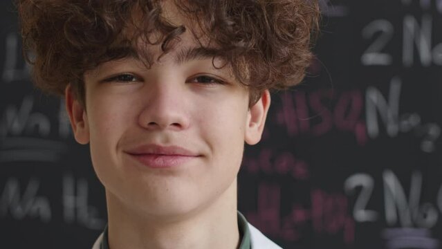 Zoom in portrait of curly haired Biracial school boy smiling at camera standing in front of blackboard with chemical formulas