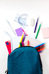Top view: a blue backpack from which stationery breaks out on a white background. Notebooks, felt-tip pens, pens, colored pencils, stickers, rulers, figured rulers, eraser. Back to School