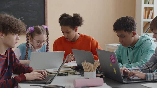 Waist up of multiracial preteen students using laptop computers and chatting while sitting together at desk during IT lesson in modern classroom