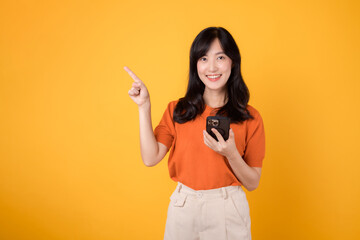 Cheerful young Asian woman in her 30s, wearing an orange shirt, points to free copy space while using smartphone on yellow background. new app concept.