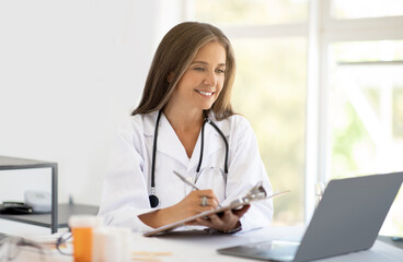 Cheerful adult caucasian nutritionist doctor in white coat at table makes notes in clipboard, looks at laptop