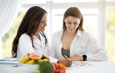 Cheerful adult caucasian nutritionist doctor in white coat makes notes of weight loss plan to young...