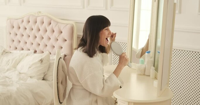 Cheerful woman sits at vanity table, embracing the joy of singing as she uses a comb as a makeshift microphone. With a beaming smile and twinkle in eyes, she playfully lip-syncs to her favorite tune.