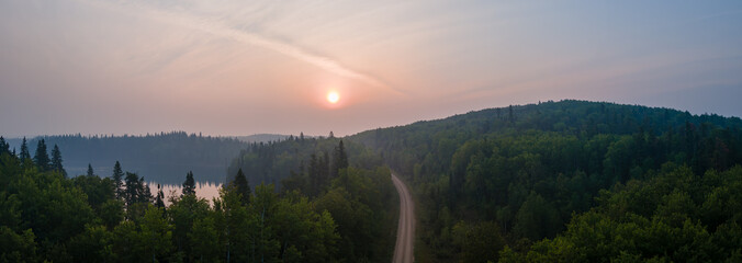 Panoramic aerial view of a green northern forest and a dirt road under a smoky orange colored sky. The corner of a calm lake occurs on the left side.
