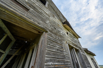 Looking up at a steep angle at a dilapidated old wooden farmhouse. The sky is filled with white to bluish clouds.
