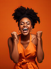 Positive contented curly haired African-American girl raising her fists, celebrating an achievement triumph against orange background