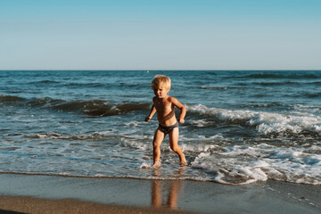 Fototapeta na wymiar little blond boy 3 years old walks on the water by the sea