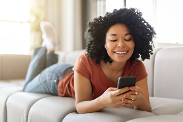Smiling Young Black Woman Relaxing With Smartphone On Couch At Home