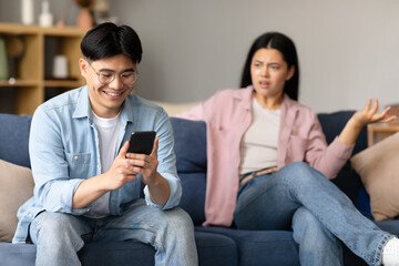 Asian Husband Texting On Smartphone Ignoring His Displeased Wife Indoor