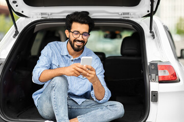 Happy indian guy sitting in open car trunk, using smartphone