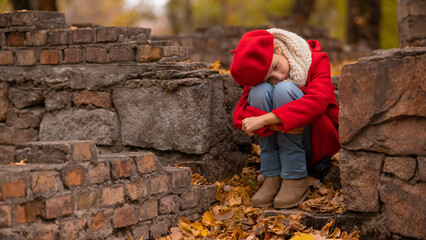 Sad caucasian girl in a red coat and beret sits on a brick wall on a walk in autumn. 