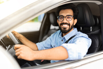 Happy eastern guy driving white auto, smiling at camera