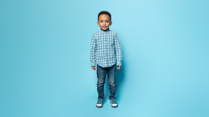 Cheerful little black boy in casual standing isolated over blue studio background, full length,...