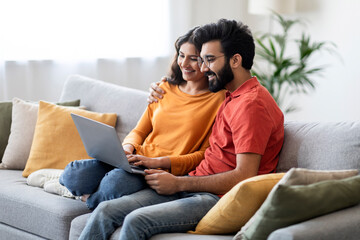 Millennial Happy Indian Couple Relaxing With Laptop On Couch At Home