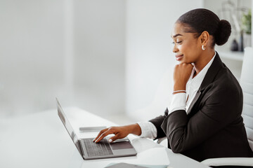 Confident black businesswoman looking at laptop and smiling, sitting at workplace in office, side...