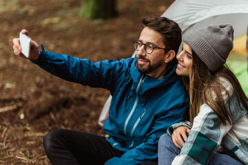 Cheerful young caucasian couple in jackets enjoy travel vacation with tent, make selfie on smartphone - Powered by Adobe