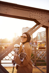 A hijab woman in stylish sunglasses and an elegant French outfit, walking through the city at sunset, carrying a bouquet, bread, and newspaper, radiating a sense of cultural charm and serenity