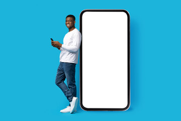 Positive young black guy standing by huge phone, using smartphone