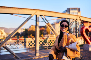 A hijab woman in stylish sunglasses and an elegant French outfit, walking through the city at sunset, carrying a bouquet, bread, and newspaper, radiating a sense of cultural charm and serenity