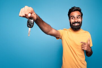 Excited indian man driver holding in hand new car key, pointing at it and smiling isolated on blue...