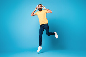 Full length shot of excited middle aged indian man wearing headset listening to music, jumping over blue background