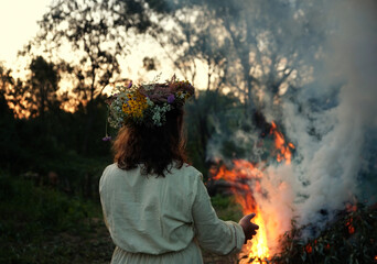 Girl in floral wreath near campfire in forest, dark nature background. rear view. Magic ceremonial,...