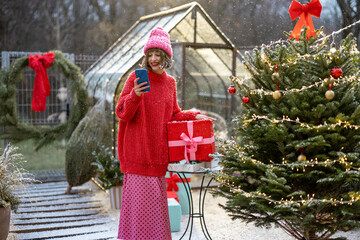 Woman in red sweater and hat using mobile phone while standing with a gift box near Christmas tree at beautifully decorated backyard during a snow fall. Concept of winter holidays and communication