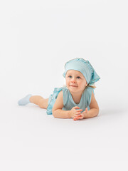 A beautiful 2-year-old toddler girl in a blue dress and a scarf lies on her stomach on the floor and looks up attentively on a white background. Photo