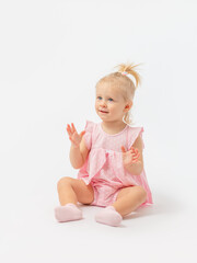 A beautiful Caucasian girl 1-2 years old in a pink dress is sitting on the floor watching and clapping her hands on a white background. Copy space.