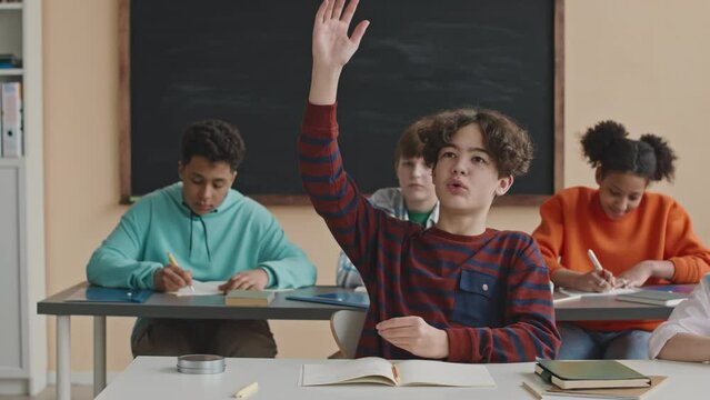 Waist up of Biracial preteen schoolboy at desk raising his hand to ask teacher question while having lesson in bright modern classroom