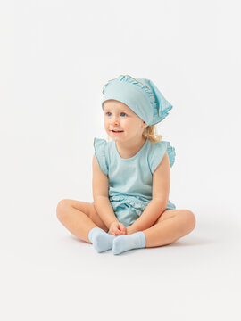 A Caucasian 2-year-old toddler girl in a blue dress and a kerchief is sitting on the floor and attentively looking at something on a white background.