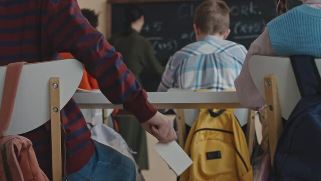 Back view of schoolboy passing cheat sheet to his classmate while having Math lesson in modern class