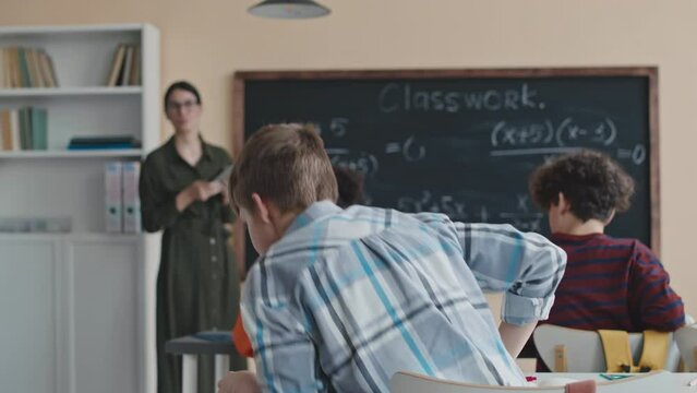 Back view slowmo of Caucasian schoolboy in checkered shirt raising his hand, standing up from his place and going to blackboard to solve difficult Math equation