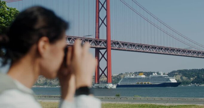 A woman photographs a river with a red bridge over it and a motor ship passing through it. Women's face is blurred