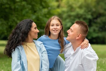 Young of Mixed Race Couple and their Daughter had a Day Off and a Picnic in the City Park. Mom and Dad Look at Their Child with Love. Girl Hugs her Parents and Looks, Smiling Brightly at the Camera.