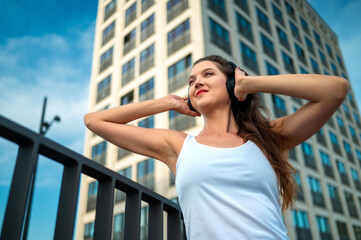 Low angle perspective of an attractive young woman in a white tank top against a skycraper listening to music on headphones