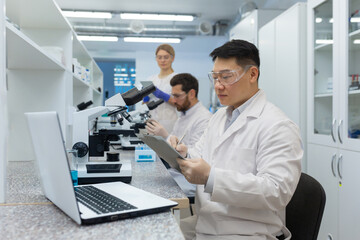 A team of scientists and doctors work in the laboratory. Asian man writing results and sitting at laptop, male and female colleagues examining under microscope.