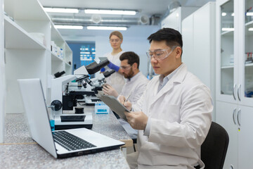 Fototapeta premium A team of scientists and doctors work in the laboratory. Asian man writing results and sitting at laptop, male and female colleagues examining under microscope.