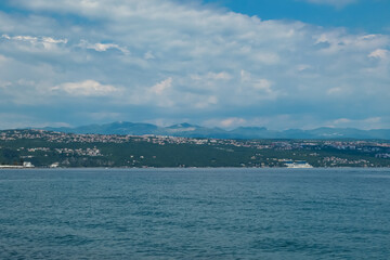 Fototapeta na wymiar A panoramic view of the Croatian shore. There are some towns located on the shore of the Mediterranean Sea. Calm surface of the sea. Stony beach. Green hills in the back. Sunny day with puffy clouds