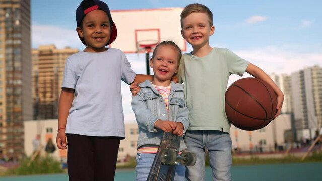 Children Multiethnic diverse are standing on playground, hugging and laughing with happiness. Caucasian children hug dark-skinned boy and laugh holding a skateboard and basketball in their hands.